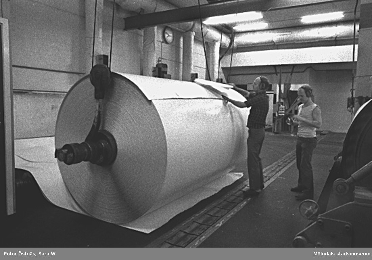 Tambourn är lyft ur kartongmaskinen och Denny Andersson och Jan Callesen tar prov på kartongen för att kontrollera kvalitet och tjocklek, Byggnad 6, våning 2. 
Bilden ingår i serie från produktion och interiör på pappersindustrin Papyrus, 1980-tal.