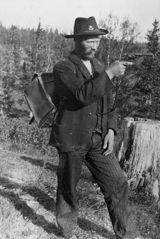 Lantbrevbärare Martin Jönsson iklädd privat hatt försedd med postemblem, siktar med revolvern. På ryggen har han en ränsel.