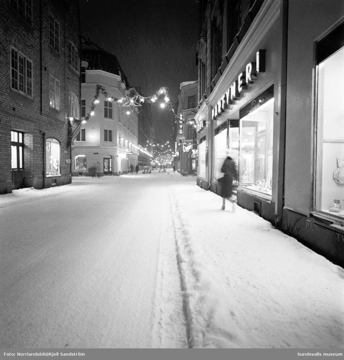 Kvällsbild från Nybrogatan i julbelysning. Två glada herrar bär hem sina julklappsinköp.
