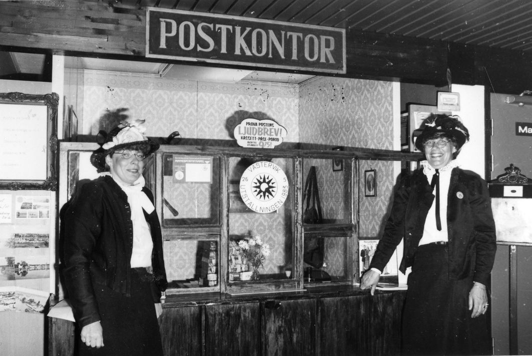Inga-Britt Södergren och Agneta Thörnberg utklädda till 1870-talets postkassörskor står framför ett postkontor från samma tid på mässan i Västervik ca 1983.