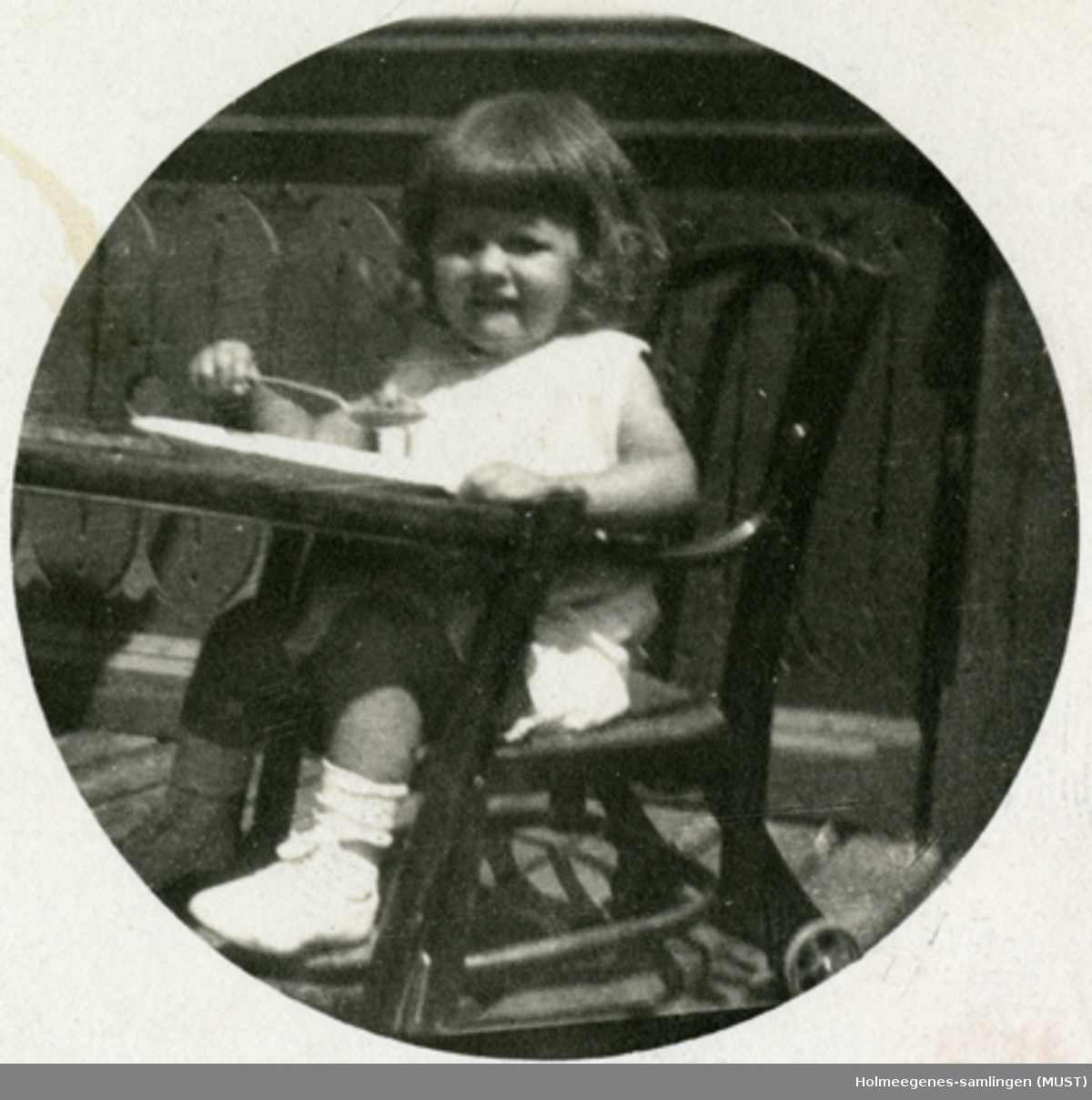 En liten jente i en barnestol, utendørs. Positiv kopiert på postkort-papir. Postkortet er påskrevet og poststemplet Wien august 1922. Jenta på bildet kalles her "Lillemor".