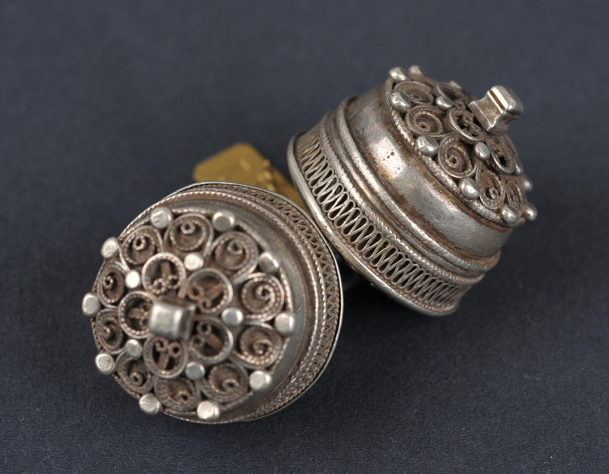 To knappar som er hekta saman med ein liten ring. Knappane har 10-dela filigransroser med 5-dela roser oppå. Kolteknapp.