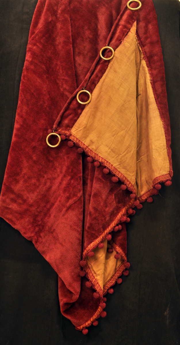 Tykk, mørkerød gardin med gylte opphengsringer, bånd langs kant med mørkerød og gulbrun tråd.