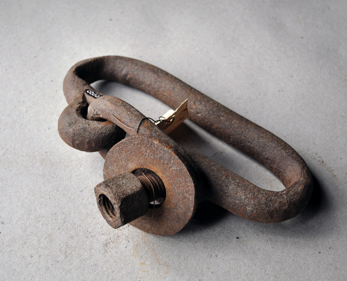 Håndmidd sjakkel av jern. Oval form med lås midt på langsiden. Låsen består av en bøyle som låses over en pinne med mutter og skive.