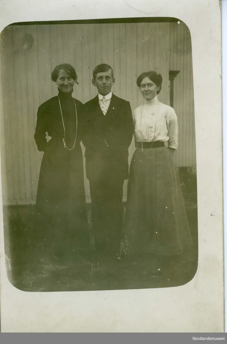 Postkort med fotografi av tre mennesker; to kvinner og en mann i midten. Kvinnen til høyre har svart kjole og langt halskjede. Kvinnen til høyre har grått skjørt og hvit bluse med svart belte i livet. Mannen i midten har mørk dress, hvit skjorte og slips. Hvit trevegg i bakgrunnen.

Bakside blank.