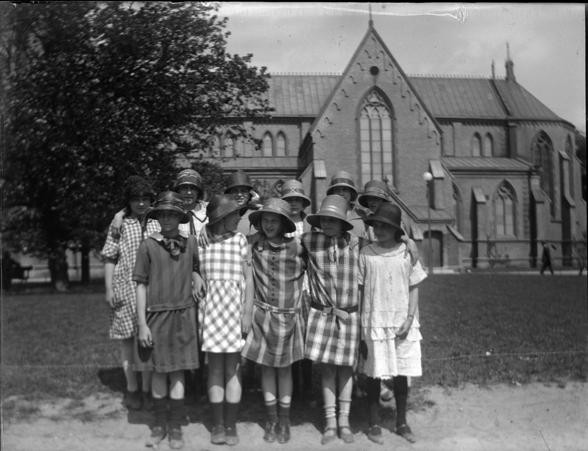 'Bildtext: ''Skolbarn på Hagaplan.'' :: Gruppbild med 11 hatt och klänningsklädda flickor samlade på en plan framför byggnad. ::  :: Ingår i serie med fotnr. 5224:1-13.'