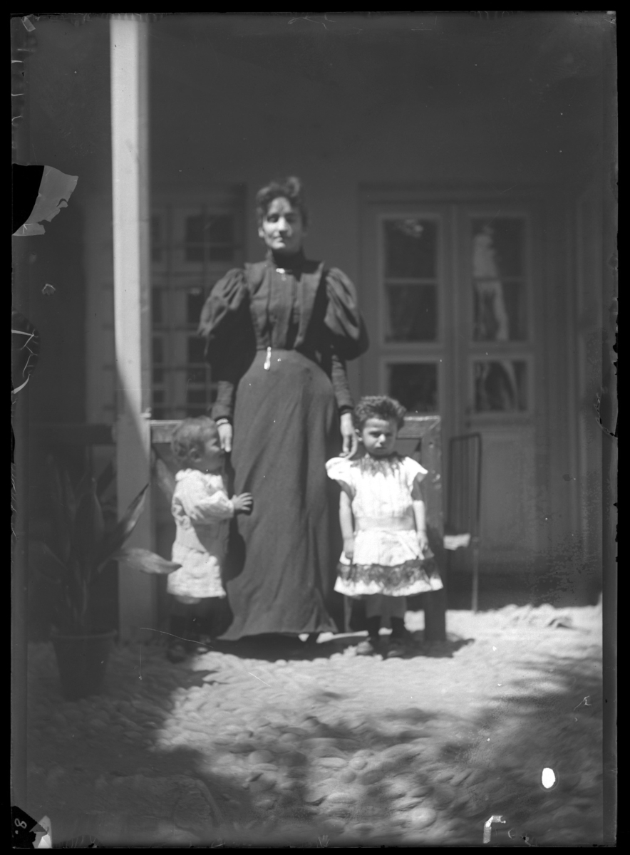 '1 kvinna med 2 små barn stående framför glasad dörr till hus. Kläder. Klädsel. Kullersten. Kruka. ::  :: Ingår i serie med fotonr. 5277:1-19. Se även fotonr. 5269-5292 med bilder från Stuxbergs resa till Kaukasien.'