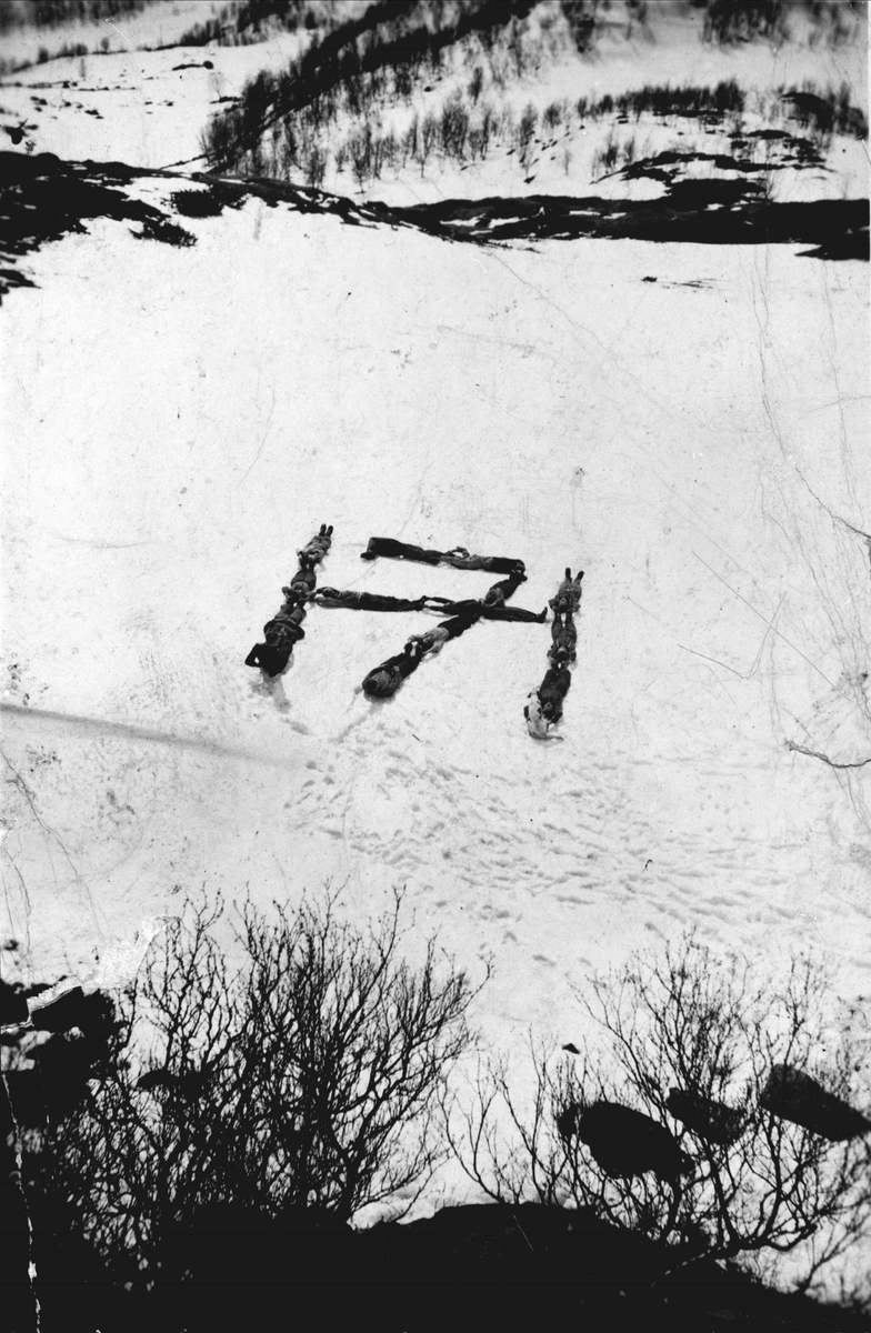 Tretten menn og en hund lager Haakon den syvendes monogram i snøen på Blåhatten.