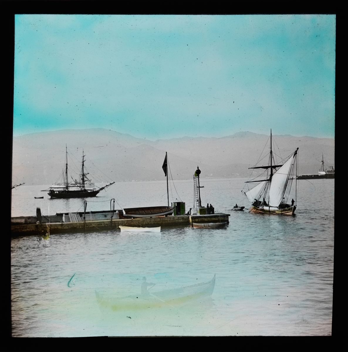"Smyrna." (Bildetekst fra album) En kai/brygge. Mange seilbåter ute på vannet. Håndkolorert utsnitt.