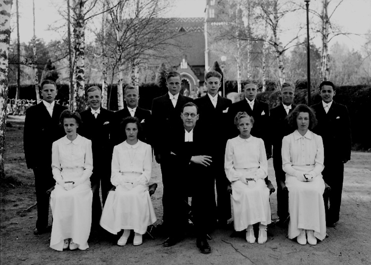 12 konfirmander, 4 flickor, 8 pojkar och kyrkoherde Eneström.
Längbro kyrka i bakgrunden.