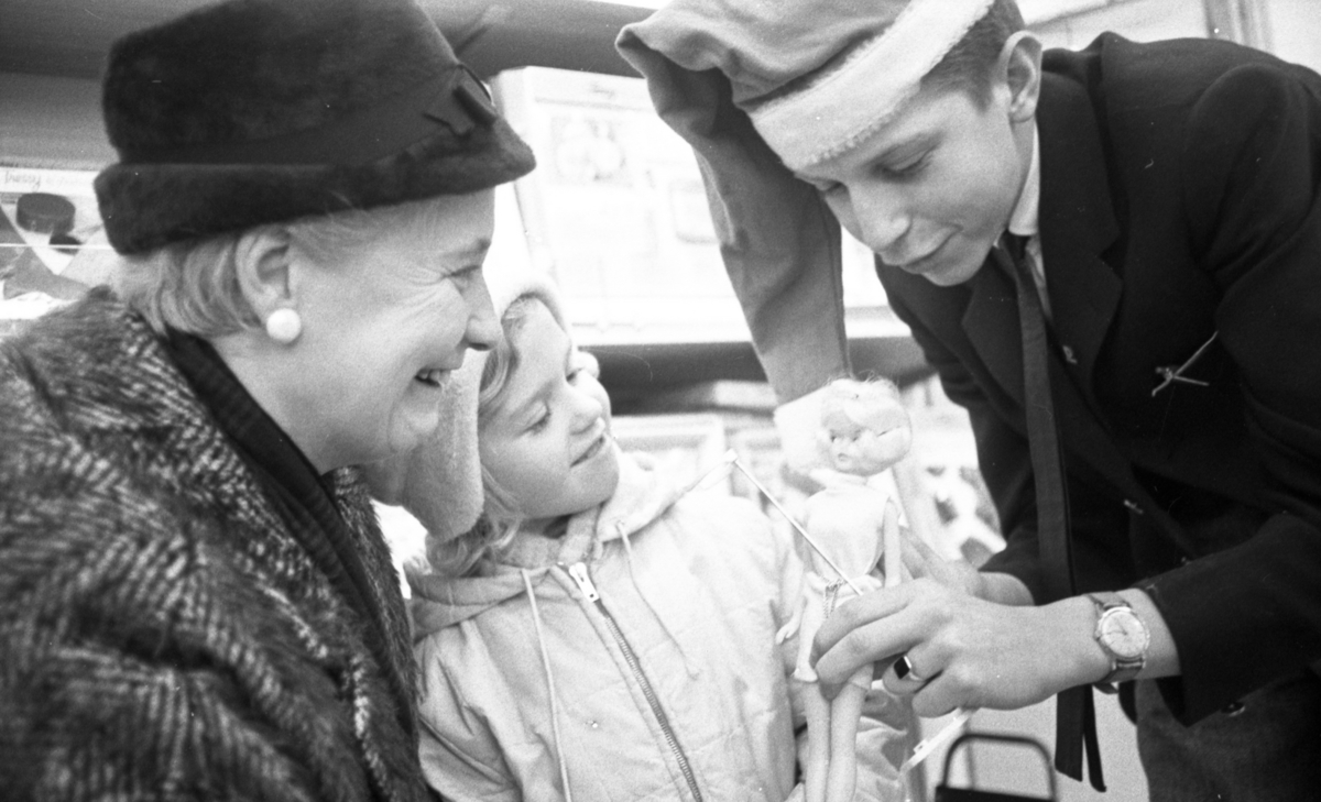 Julköprush, 7 december 1965

En ung man i tomteluva och svart kostym visar en docka  för en liten flicka och en äldre dam.