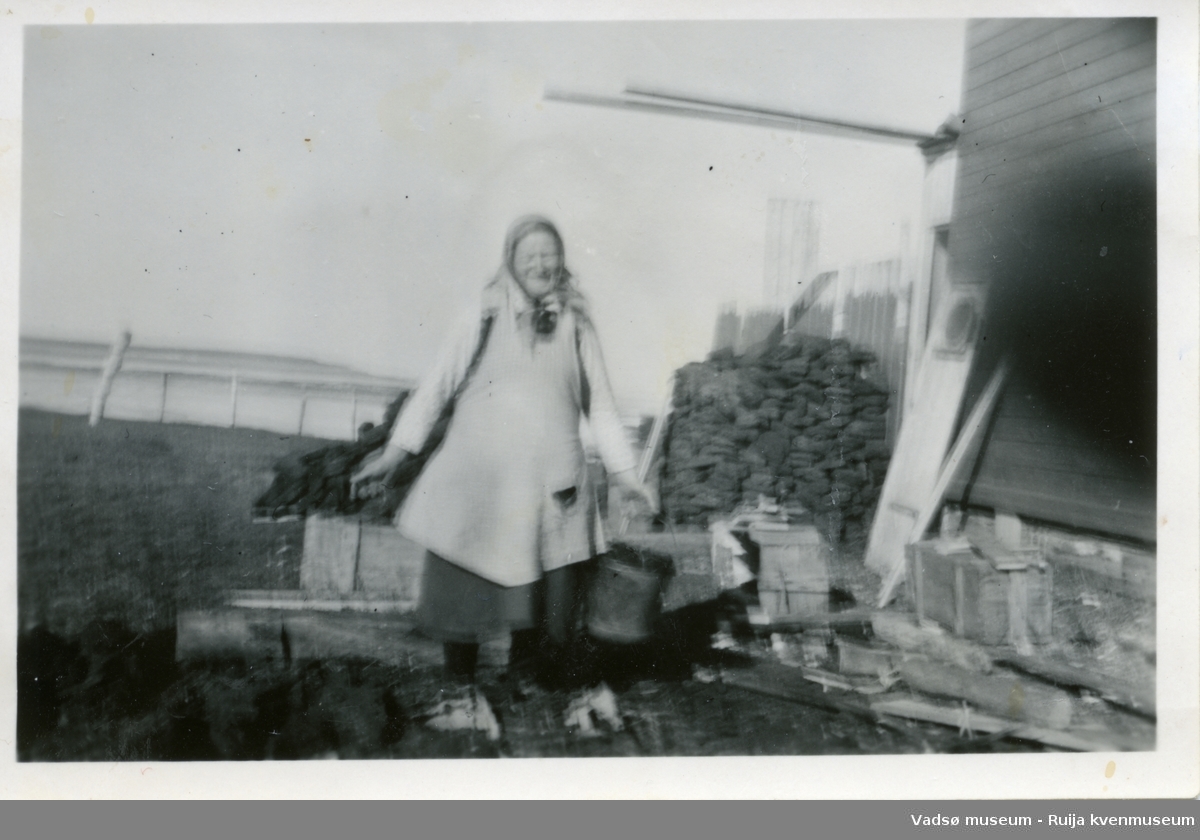 Grete Julkunen avbildet på baksiden av huset mens hun bærer en bøtte torv fra torvstabelen i bakgrunnen.