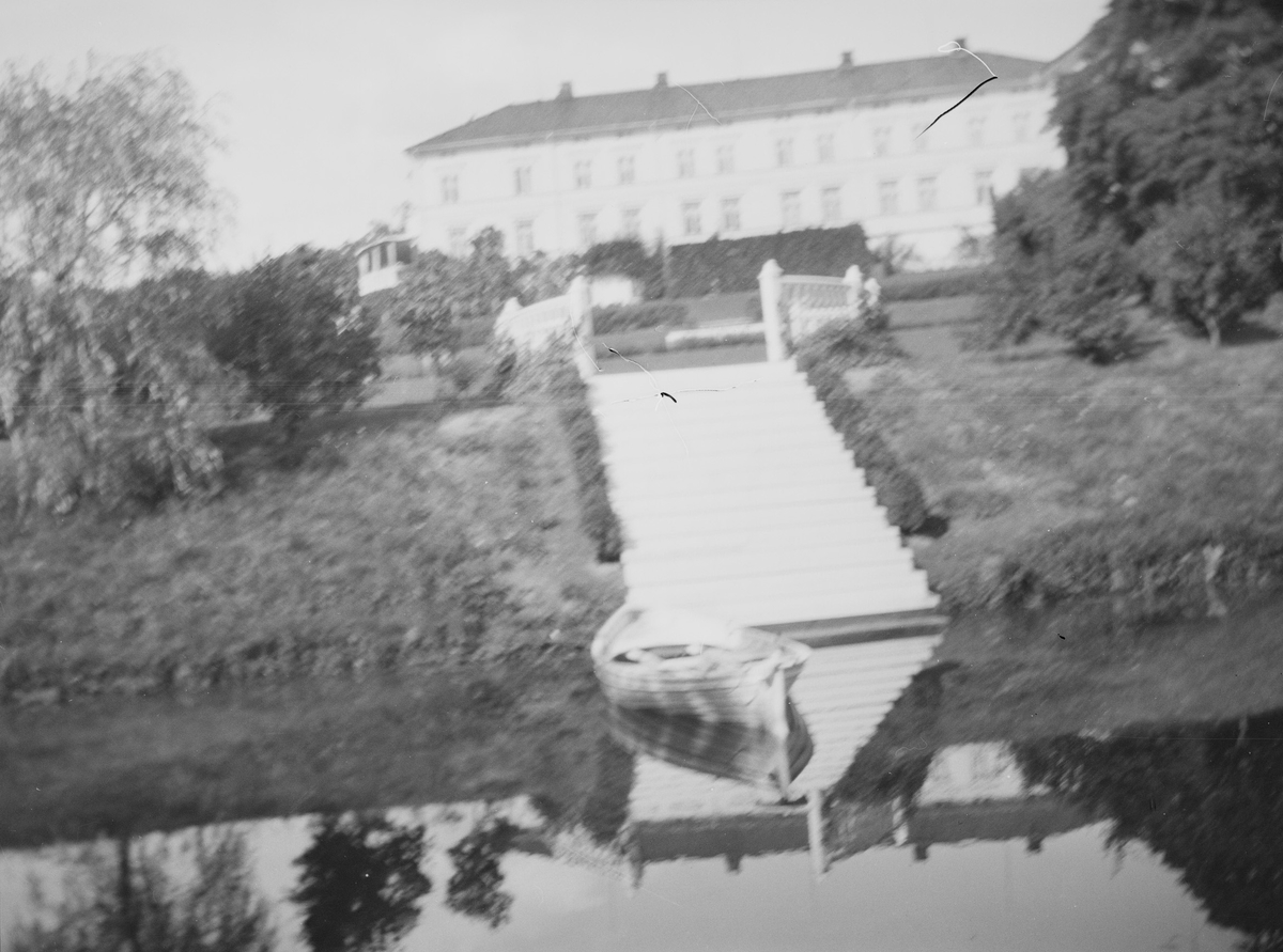 En robåt på dammen i hageanlegget på Linderud Gård, om sommeren. I bakgrunnen sees hekker, trær samt hovedbygningen.