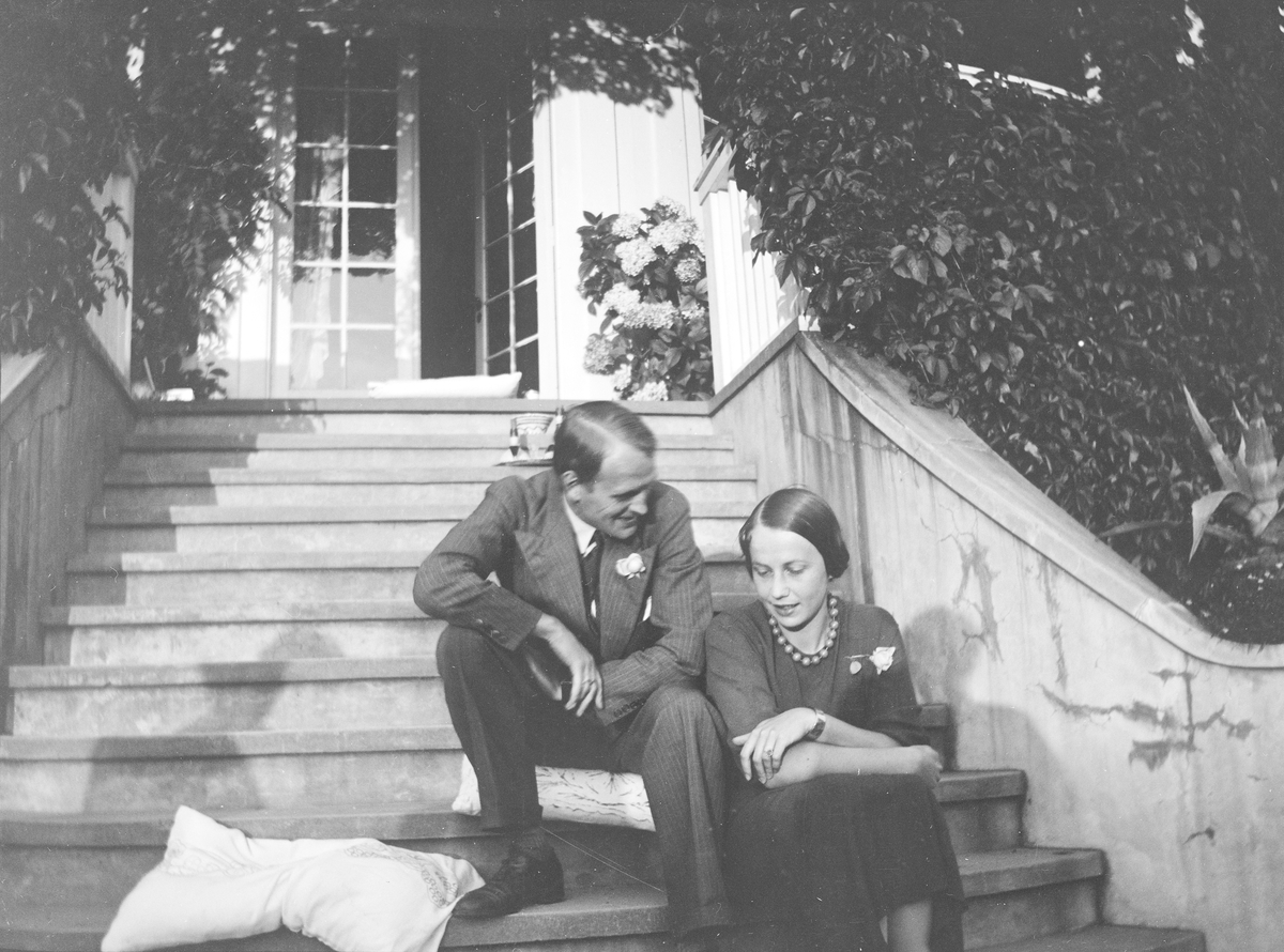 En kvinne og en mann sitter på trappen opp til verandaen fra den "Victorianske hagen"på Linderud Gård. De har tatt med puter som de sitter på. Verandaen er dekket av klatreplanter.