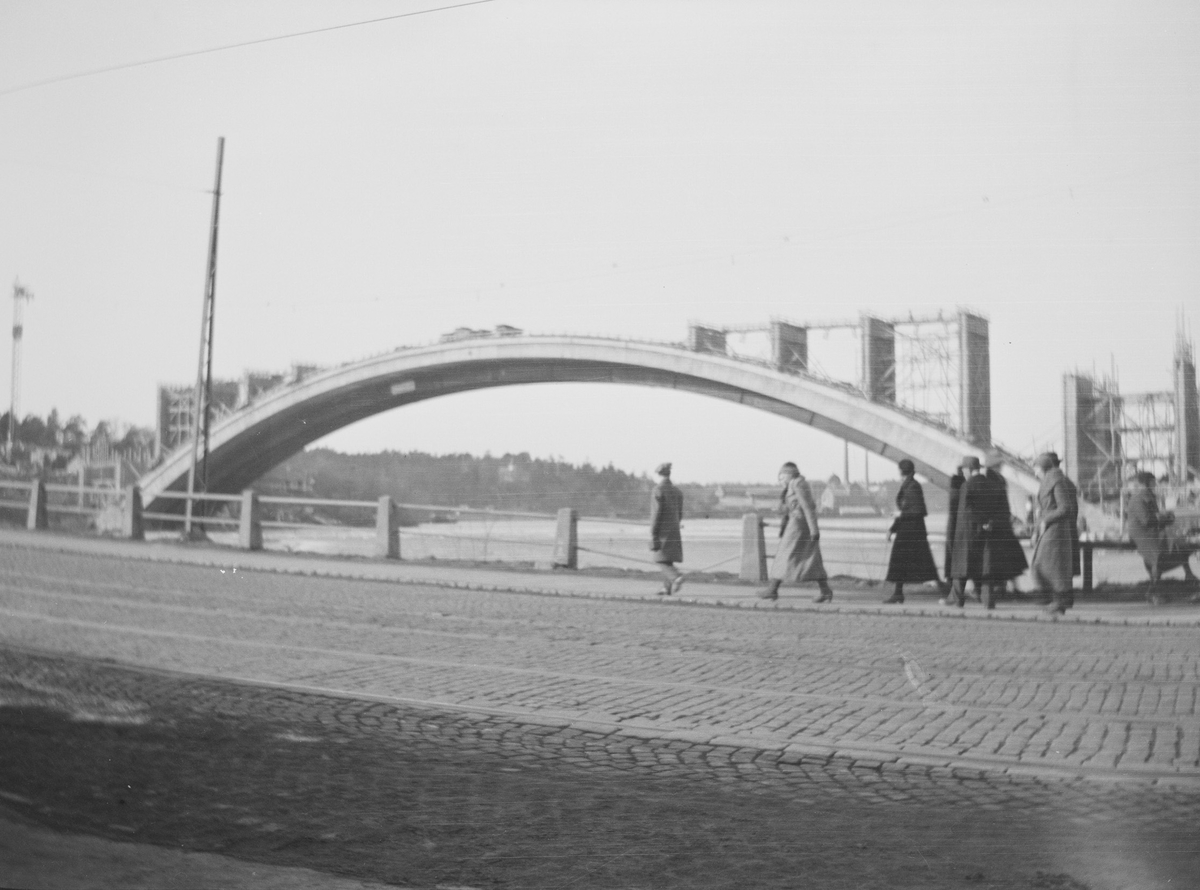 Menn og kvinner kledd i vinterklær på vei fra Gamla Stan mot Söder. Söder sees i bakgrunnen. Dette er i området hvor Slussen ligger og kanskje en eldre utgave av Skeppholmsbron.