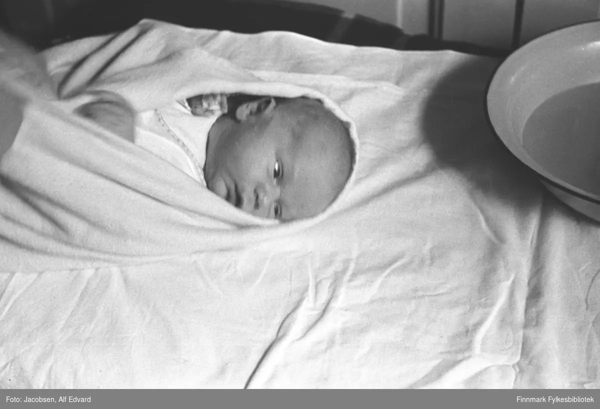 Arne Jacobsen som nyfødt baby. Spedbarnet er inntulllet i et lyst/hvitt laken eller teppe og har en hvit drakt på seg. Et vaskefat som er halvfullt av vann står på bordet der barnet ligger. En liten del av veggen ses i øverkant av bildet og viser lyst, stående panel.