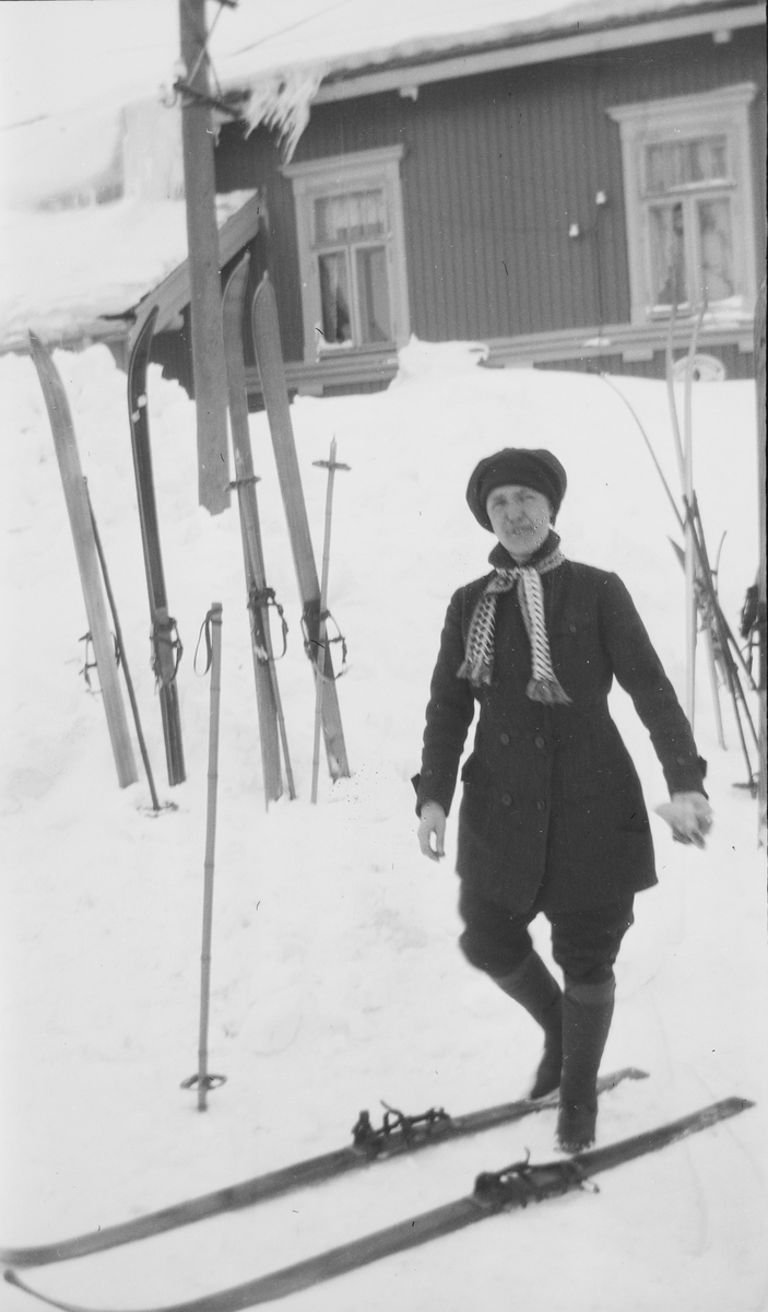 En kvinne står foran Haugastøl Stasjon og Turisthotel. Enten er hun i ferd med å ta på seg ski eller hun har akkurat tatt dem av. Mange par ski og staver står plantet i snøen.