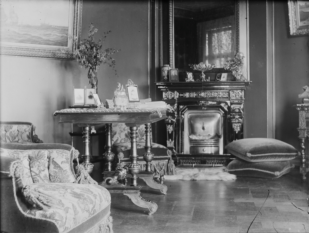 Fotografi av stue med kamin med speil over i hjørnet, sofa og stoler med detaljer som "klunker", bord med utskjæringer, fotografier i rammer på bord og kamin, puter og duker, malerier på veggene og en blomstervase med avskårne blomster på bordet.