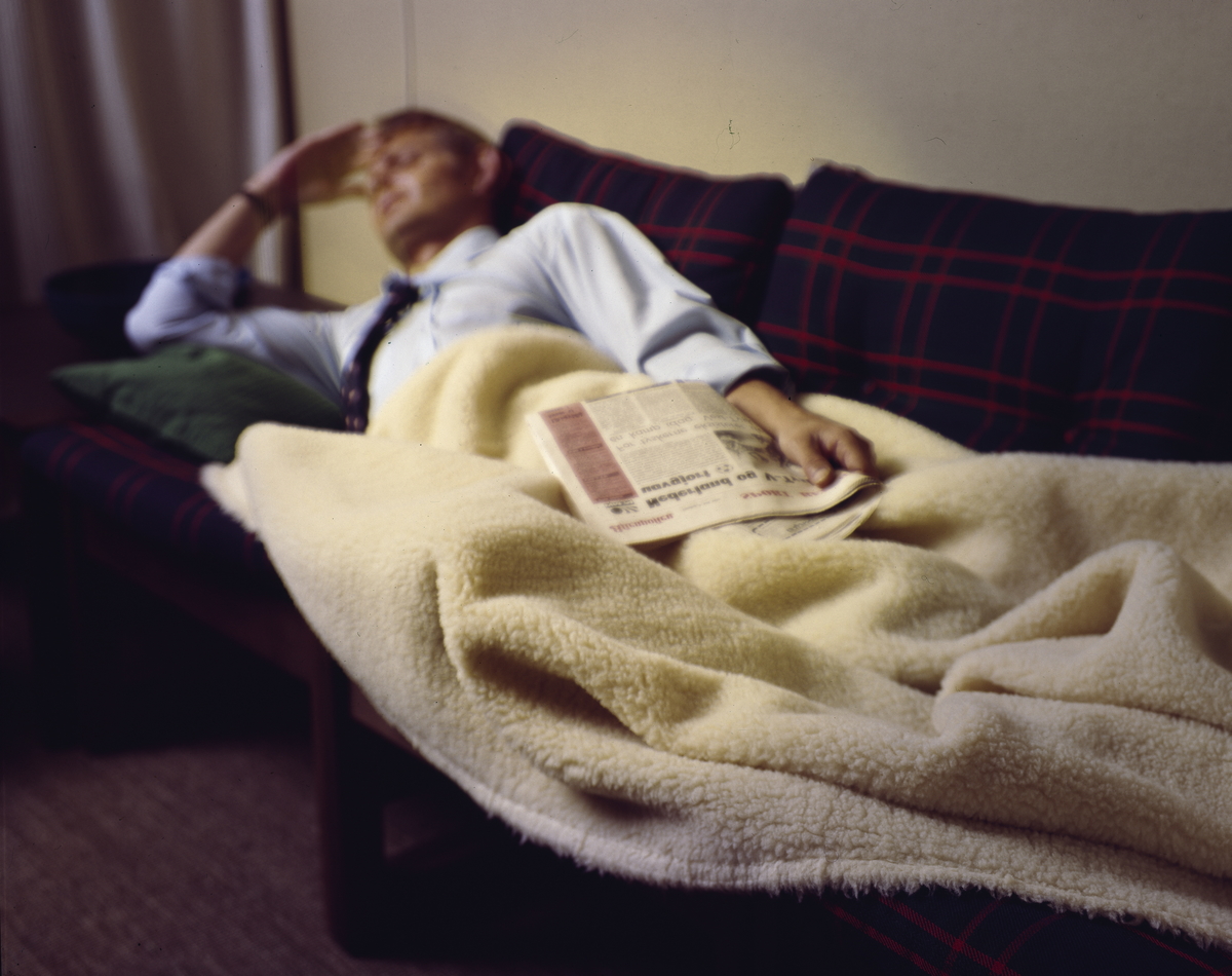 Reklame- eller illustrasjonsfoto av en mann som ligger på en sofa med avisa i hånda og et pledd over seg. .