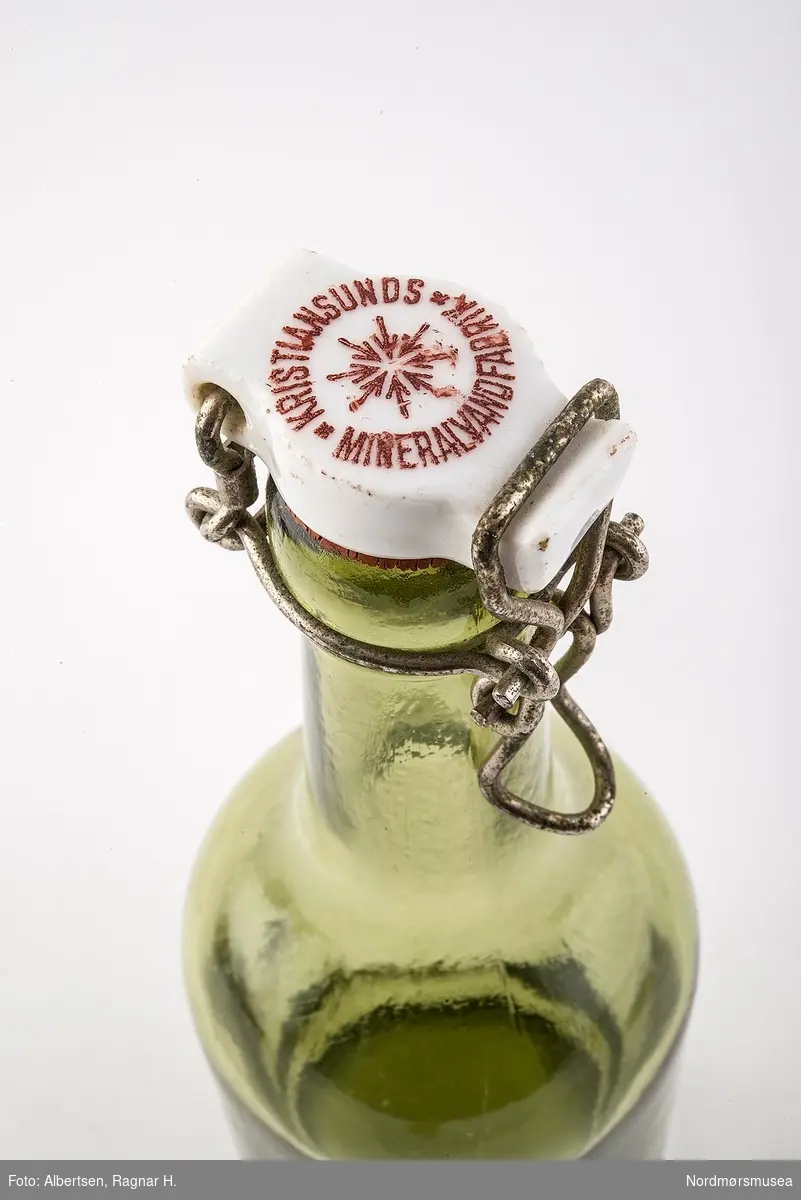 Rund grønn glassflaske med slake skuldre. Toppen er laget av steingods med korkdelen som stikkes ned i flasken. Toppa holdes med metall-hekting (svingtopp).