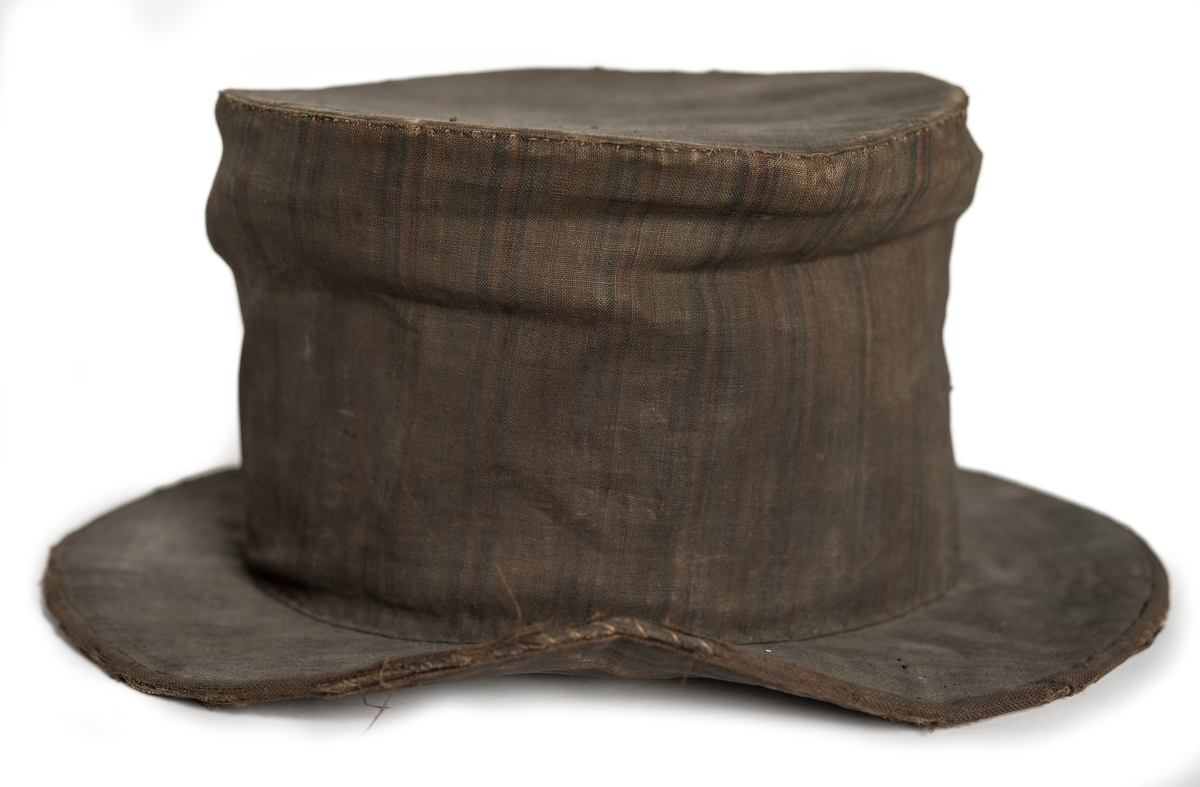 Hög hatt med brätten, överdragen med mörkt randigt linnetyg i fyra färger, övervägande brunt. Svettrem längs insidan av svart läder. Fodrad med vit lärft. Skullen uppbyggd av halm.