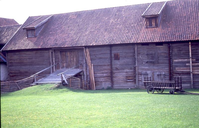 Bildet viser en eldre laftet låve. det er grønt gress og låven er stor. Den henger sammen med fjøset og stallen i endene.