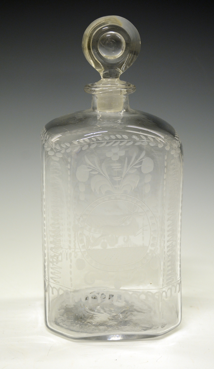 Glasskaraffel i klart glass - firkantet form, med avbrutte hjørner (skrå), slik at flasken eg. har 8 kanter. Gravering på alle sider - midt på begge de store flatsidene er det gravert et dyr (hjort eller rådyr), ellers vanlige ranker, blomster og blader.
Flasken har glasspropp - virker original. Ingen skader.

Stor glassflaske av typen kantineflaske. Gravert. Usikker produsent - kan det være Gjøvik eller Hurdal? 1790-1820 ca.