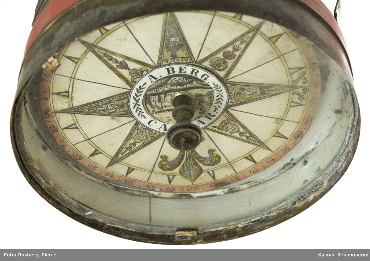 KLM 44862. Kompass, skeppskompass av metall, glas. Huset av metall översidan krönt av en kronliknade dekoration av metallband. Del av huset målat rött. En så kallad skvaller-kompass eller enligt Ekbohrn, Nautisk Ordbok, 1840: häng-kompass, hänger under skylight i kajutan sålunda att på den kan ses, äfven för kontroll, huru rorgängaren styr eller ligger an. Kompassen signerad, A. Berg. Calmar.