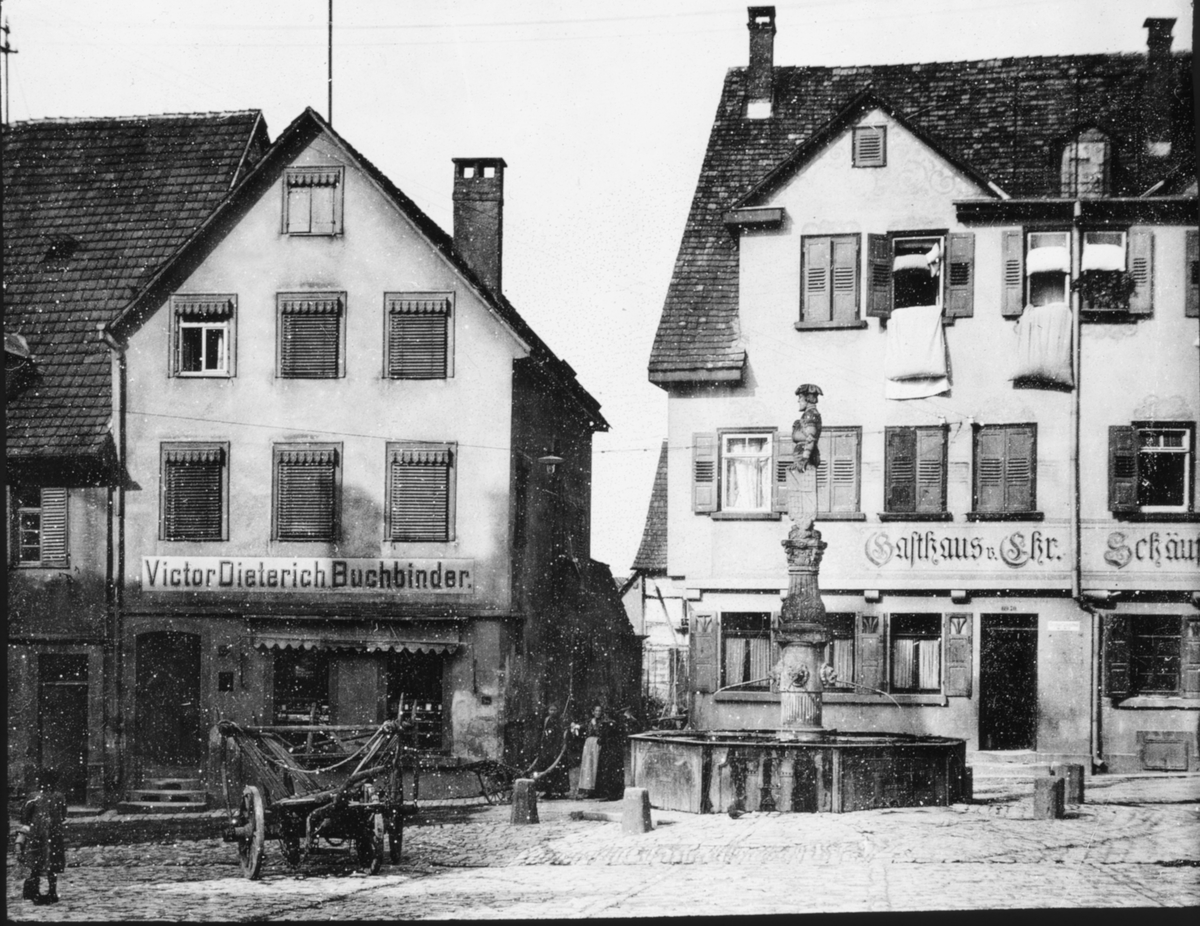 Skioptikonbild med motiv av fontän på torg vid bokbinderi i Besigheim.
Bilden har förvarats i kartong märkt: Resan 1907. Besigheim 8. 29