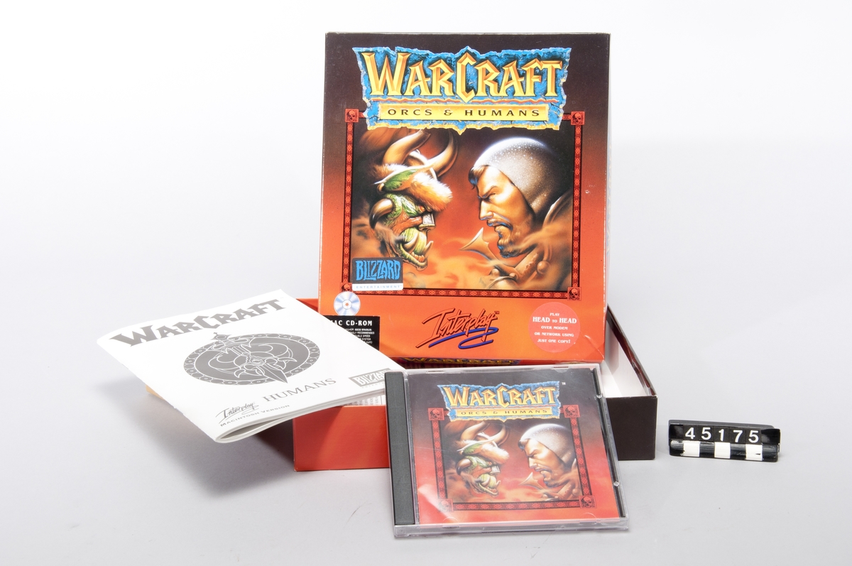 Röd box av papp med titeln "Warcraft Orcs & Humans", innehållande en CD-ROMskiva med spelet samt en tryckt manual på engelska. Spelet är för Macintoshdatorer.