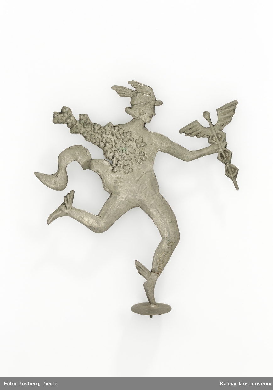 KLM 28228:9. Prydnadsfigur, till blomsterdekoration. Av metall, tenn. Figur föreställande en man, guden Hermes i grekisk mytologi. Han har hjälm och skor med vingar på och håller i en spira med vingar. Hans bål och ena arm är inlindande i blommor. Hans kläder rör sig i vinden. Han står på ett ben, benet står på en rund platta med skruv och mutter på undersidan.