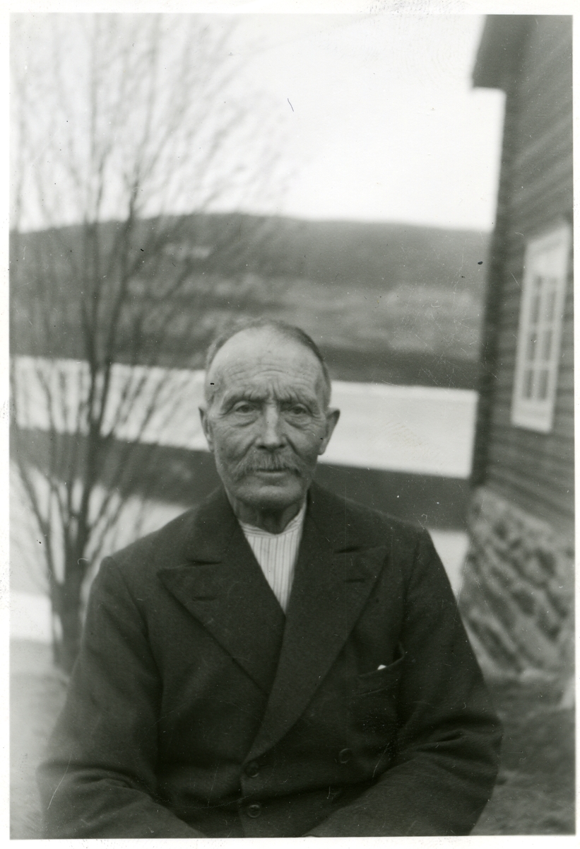 Portrett av en mann sittende foran et hus. Mannen er iført en mørk dressjakke og en stripete skjorte.