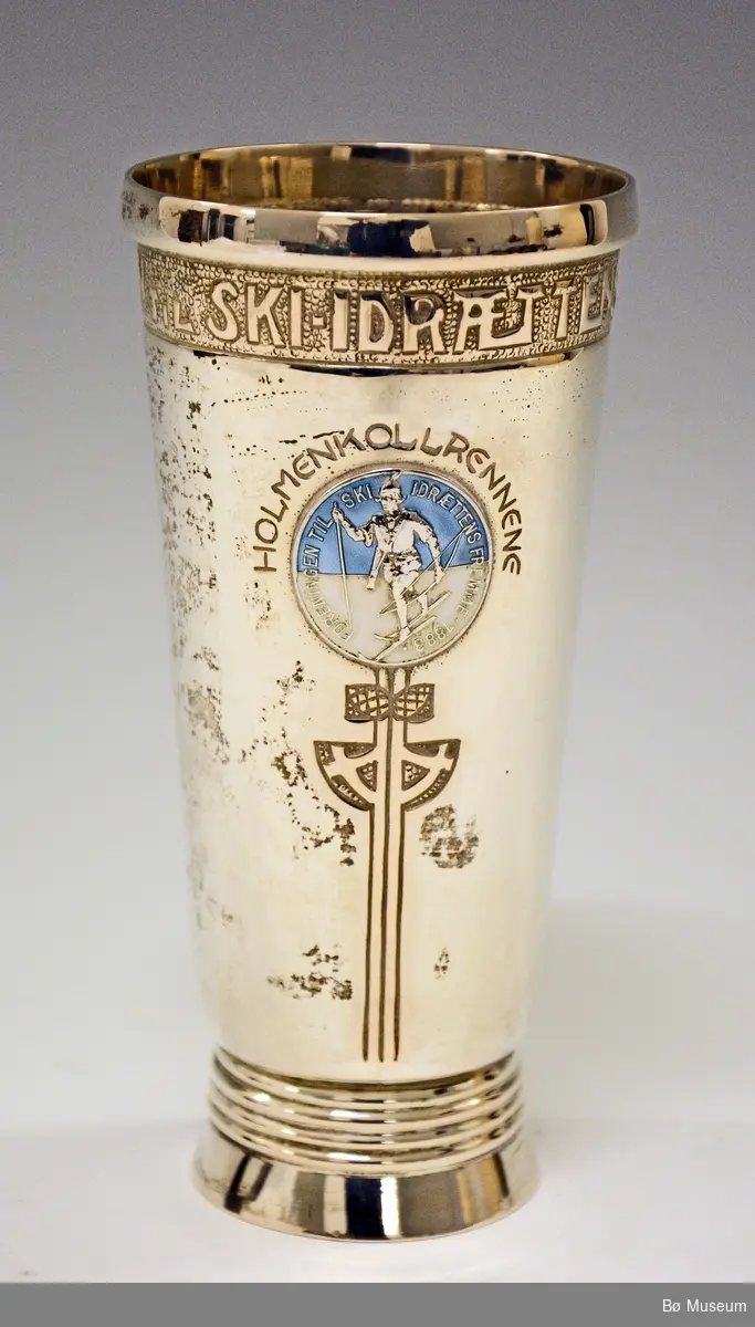 Sølvpokal med merke i blått foran. Merket har følgende innskrift: 
"Foreningen til Ski-idrættens Fremme 1883"
Rundt dette merket står:
"Holmenkollrennene" (u årstall)