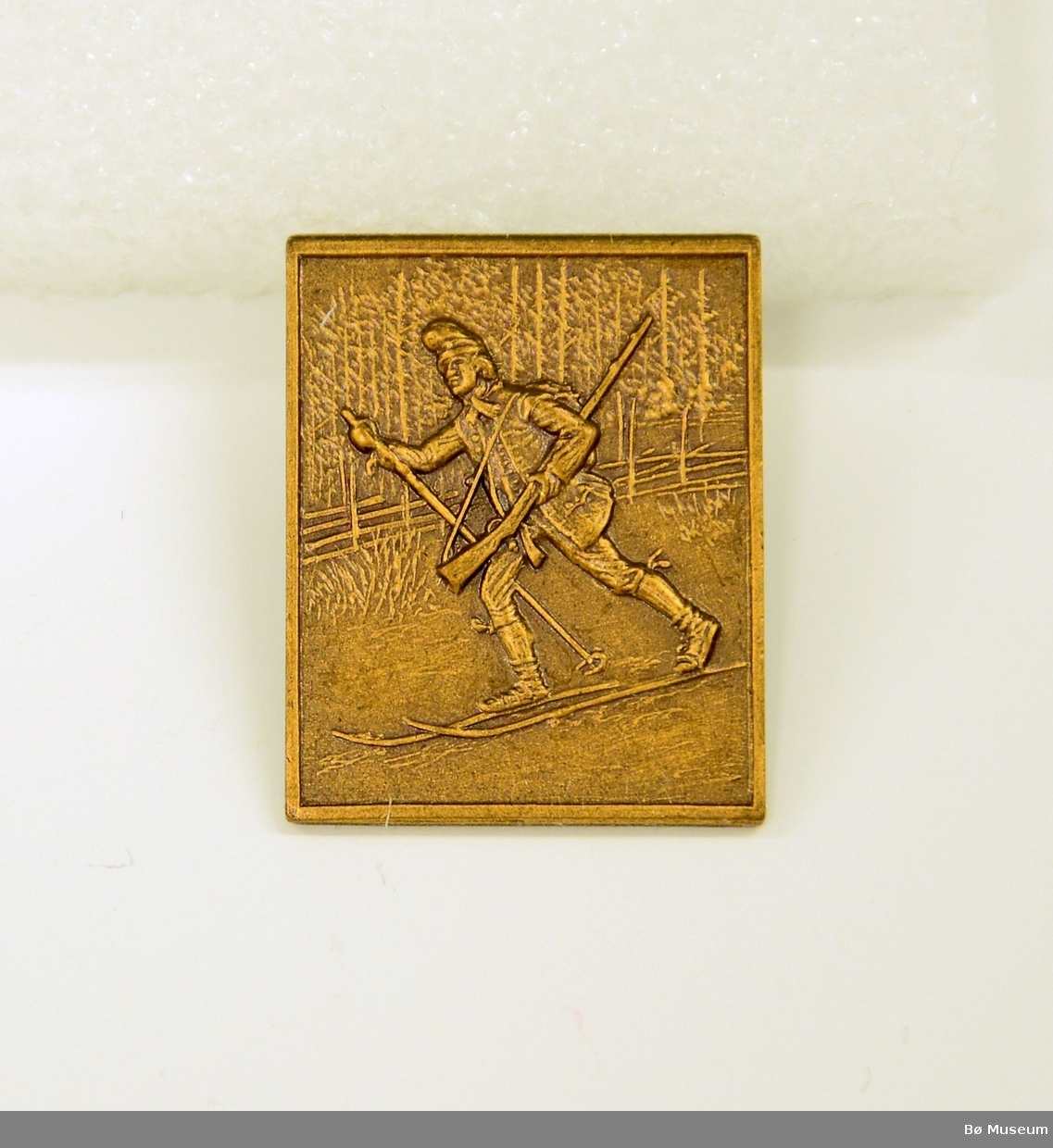Pin - uten innskrift:
I messing/kobber - bilde av en mann på ski med gevær - Jo Gjende? Med nål bakpå.