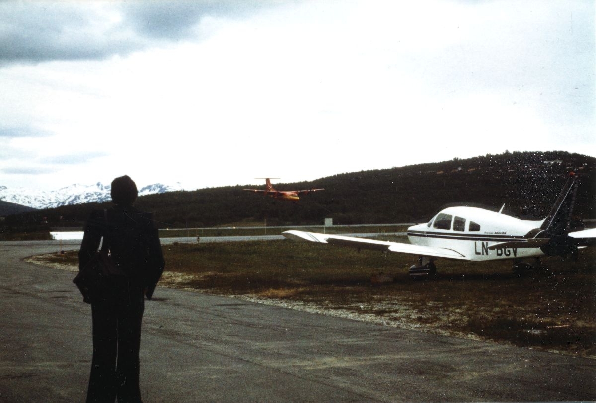 Lufthavn/Flyplass. Tromsø, Langnes. En person ser landingen til et fly, DHC-7 (Dash7) i flyfabrikken Bombardiers farger. På bakken står et fly av typen Piper Cherokee Archer parkert.