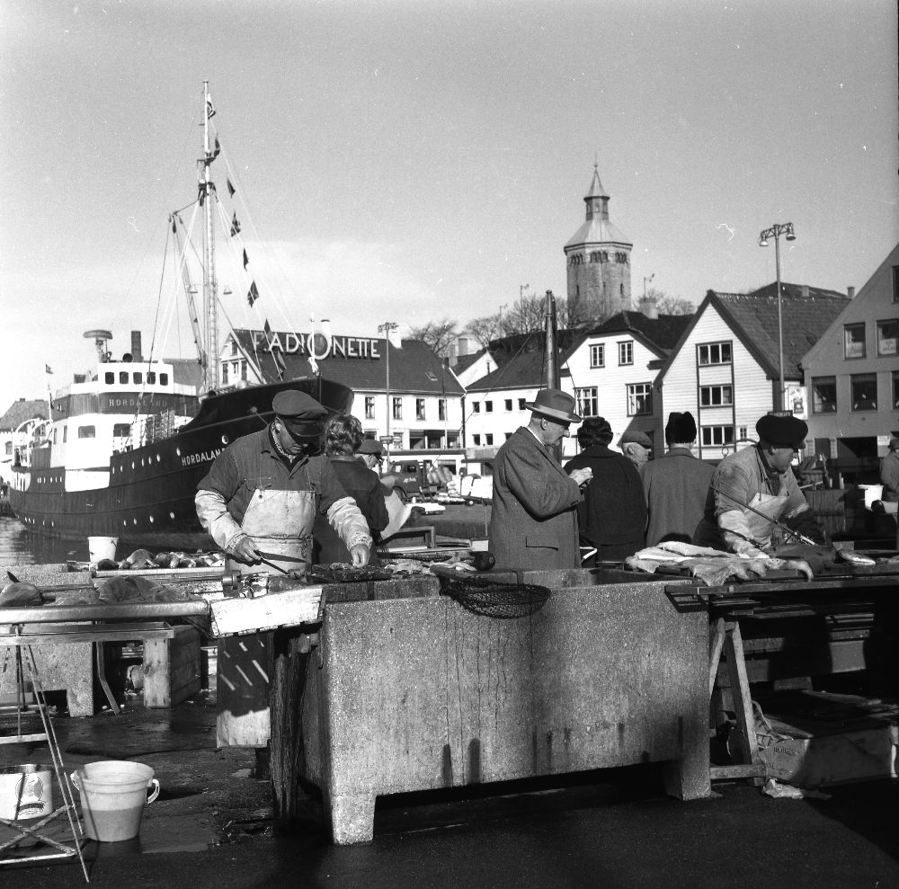 Landskap. Fisketorget i Bergen. Flere personer, selgere og kjøpere. Fisken ligger klar for kundenes middag.