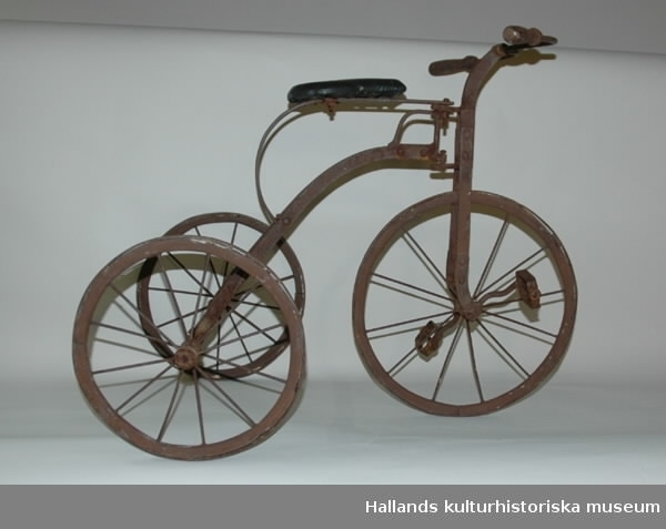 Trehjulig barncykel med metallskodda trähjul. Sadel av galon (i efterhand ditsatt). 