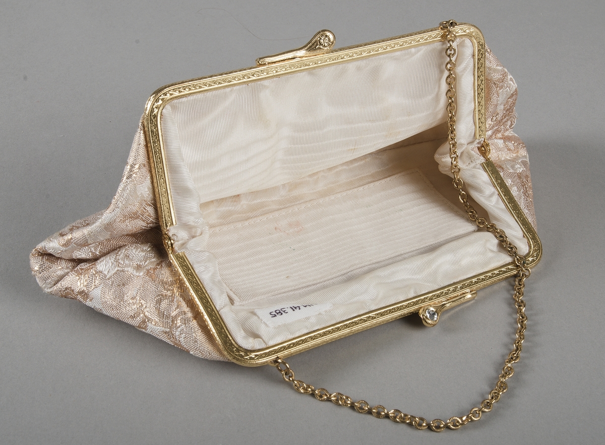 Handväska i guld, vitt och beigebrunt. Handtaget i form av guldfärgad kedja. Två vita stenar i spännet. Ett fack invändigt. 