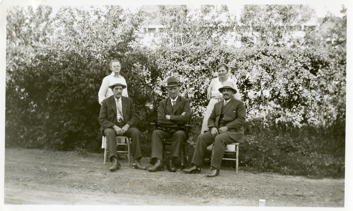 Tre menn sitter foran en hekk, og to kvinner står bak.