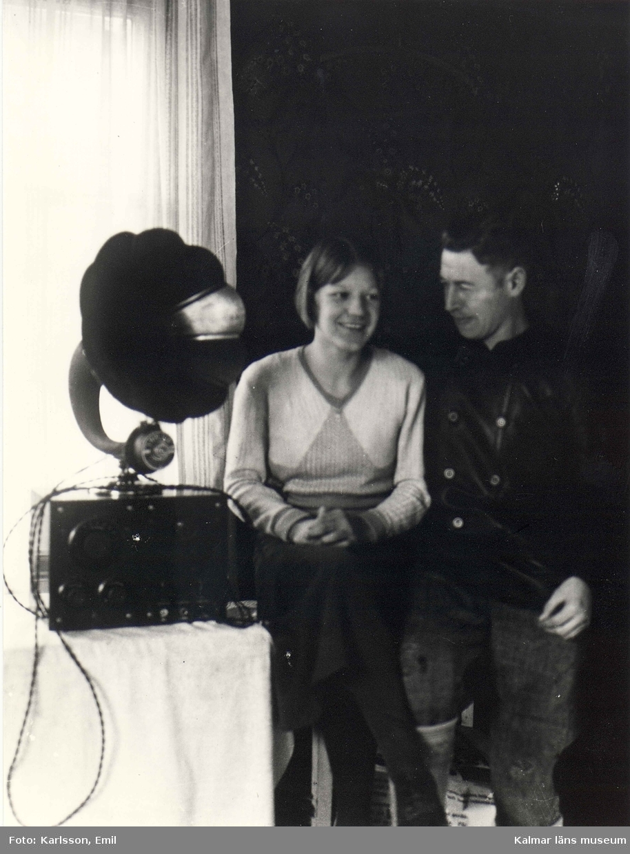 Emil Karlsson tillsammans med sin granne Greta Alexandersson, dotter till målaren Johannes Alexandersson, Mörtfors. Särskilt skall observeras den fina rundradioapparaten samt att Emil dragit för gardinen för att dämpa det infallande ljuset.