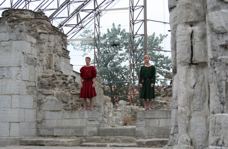Two young men in Medieval medieval costumes are standing barefoot singing in the medieval cathedral ruins.