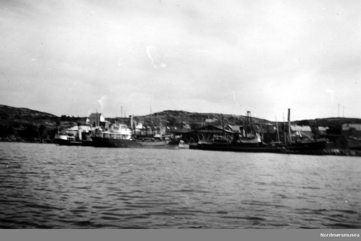 Bildet viser Storviks Mek. Verksted på Dahle ved Kristiansund. sett fra sjøsiden med båter ved kaiene og på patentslippen.
På den nye patentslippvogna klinkbygd i stål 1918, den største slippen mellom Bergen og Trondheim, ses sidetråleren S/T”Rairo” F-32-H fra Hammerfest Havfiskeselskap (senere Findus). Ved kjelkrankaia ligger tre sidetrålere, den ytterste er M/T”Nordhav 4 eller 5” til rederiet Nordhav M/T”Nordhav 4 eller 5” til rederiet Nordhavet i Kristiansund et i Kristiansund og innenfor 
S/T ”Honningsvåg” M-80-K fra Trålfiskernes Andelslag og innerst M/T”Nordhav 4 eller 5” til rederiet Nordhavet i Kristiansund. 
Den nye kjelkrana var klinkbygget i stål ca. 1945 og revet på 1970-tallet, var den kraftigste krana i Kristiansundsområdet og hadde en løfteevne på 25 tonn.
På venstre siden av piren ligger også en av byens sidetrålere S/T”Motind” M-99-K til Heinsarederiet og på høyre side av piren ligger en tankbåt (baklader) M/T”Tank 6”. Over tankbåten og helt til venstre ses bolighuset til verkstedet hvor familien Hjelmaas bodde, senere familien Paul Storvik. 
Bolighuset var et gammelt meieri i tømmer flyttet fra Straumsneset og bygd opp ca. 1910 og brant på 1980-tallet. 
Foran baugen på «Rairo» ses kjelverkstedbygningen fra 1905 og over kjelverkstedbygningen ses bolighuset til Ole og Lisbeth Kvalnesvik, senere Magnus Kvalnesvik og Ludvik Bergem.
Over mønet på maskinverkstedbyningen ses bolighuset til familiene  Alf og Knut Hjelmås. 
John Olsen Storviks bolig ses over høyre del av taket på maskinverkstedsbygningen.
Maskinverkstedbygningen med firmanavn i fronten ses midt på bildet og helt til høyre ses bolighuset til Ole Storvik, senere Karen Storvik,senere familien Schamaun.
Bildet er fra 1953-54. 
Dette er det samme motivet som bilde FAKf-100296.171652/KMb-1983-031.0215 og 
FAKf-100296.171653/KMb-1983-031.0232 og FAKf-100296.186143/KMb-1983-031.0215 og 
FAKf-100296.186170/KMb-1983-031.0231 og FAKf-100296.186171/KMb-1983-031.0232 tatt fra en annen vinkel..  alternativ:
Bildet viser Storviks Mek. Verksted sett fra sjøsiden, med båter og ved kaiene. Bildet viser sidetråleren S/T"Rairo" F-32-H fra Hammerfest Havfiskeselskap (senere Findus) på den nye patentslippvogna klinkbygget i stål fra 1918, den største slippen mellom Bergen og Trondheim, og på nordsiden av piren ligger sidetrålerne S/T"Motind" M-99-K til Heinsarederiet, ytterst og innenfor den ligger sidetråleren S/T"Ertnan" M-75-BV eller M-75-SM til Neerlandrederiet på Smøla. Til venstre bak "Motind" ses støperibygningen reist etter brannen i 1911, nedlagt ca. 1960 og revet ca. 1970, med et lagerbygg foran, og trekaia nedenfor støperiet hvor jektbåten lå fortøyd og utenfor trekaia lå en fortøyningsbøye som ble brukt ved slippsetting på såpehellingen. Til venstre for trekaia, helt inne i fjæra, lå 2-3 store runde avbarkede tømmerstokker i flomålet til bruk ved behov for store kjølemner til treskuter. Til venstre for tråleren på patentslippen ses såpehellingen og den minste slippen som hadde hjul. Såpehellingen ble bygget i 1901 og var den første slippen ved verkstedet. Til høyre på bildet utenpå hverandre ligger 3 sidetrålere ved kjelkrankaia for puss og reparasjon før neste tur til feltet. Ytterst ligger en av Nordhav-trålerne M/T"Nordhav 4 eller 5" til rederiet Nordhavet i Kristiansund. Innenfor denne ligger S/T"Honningsvåg" M-80-K fra Trålfiskernes Andelslag og innerst den andre M/T"Nordhav 4 eller 5" til rederiet Nordhavet i Kristiansund. Tankbåten (bakladeren) på sørsiden av piren er M/T"Tank 6". Bak trålerne ses den nye kjelkrana klinkbygget i stål ca. 1945 revet ca. 1970, den kraftigste krana i Kristiansundsområdet, og med en løfteevne på 25 tonn. Helt til høyre, den høye murpipa på kobberslagerverkstedet, som også inneholdt verkstedets egen elkraftsentral. På sundet utenfor trålerne ved kjelkrankaia ses "Storbøya" som ble brukt til fortøyning og enkelte ganger ved slippsetting på patentslippen. Over kobberslagerverkstedet og bakken på trålerne ses bolighuset til familien Ole Storvik, senere Karen Storvik, senere familien Schamaun, og over bakken på trålerne helt til høyre ses taket på det hvite bolighuset til familien Nils Holtan, senere familien Bastian Holtan, bak Sanitetshuset. Helt i høyre kant av bildet ses flerleilighetshuset, "Murgården" til verkstedet bygget først på 1910-tallet og revet ca. 1981. Over, og bak, skorsteinen på tankbåten ses taket på et av verkstedets bolighus og uthus, fra ca. 1910, og brant ned på 1980-tallet hvor familien Hjelmås bodde, senere familien Paul Storvik. Bolighuset var et meieribygg i tømmer flyttet fra Strømsnesset. Bygget med Storviks Mek. Verksteds navn på er maskinverkstedsbygningen bygget etter brannen i februar/mars 1911 og revet etter 1980 og til venstre, og over maskinverkstedet, ses bolighuset til familiene Alf og Knut Hjelmås. Helt til høyre over taket på maskinverkstedet ses taket på bolighuset til verkseier John Olsen Storvik, revet etter 1980, og over høyre ende av taket på John Olsen Storviks hus ses en firkantet transformatorkiosk i grå betong.

Bildet er fra 1953-54. Tekst av Peter Storvik. Fra Nordmøre museums fotosamlinger.
