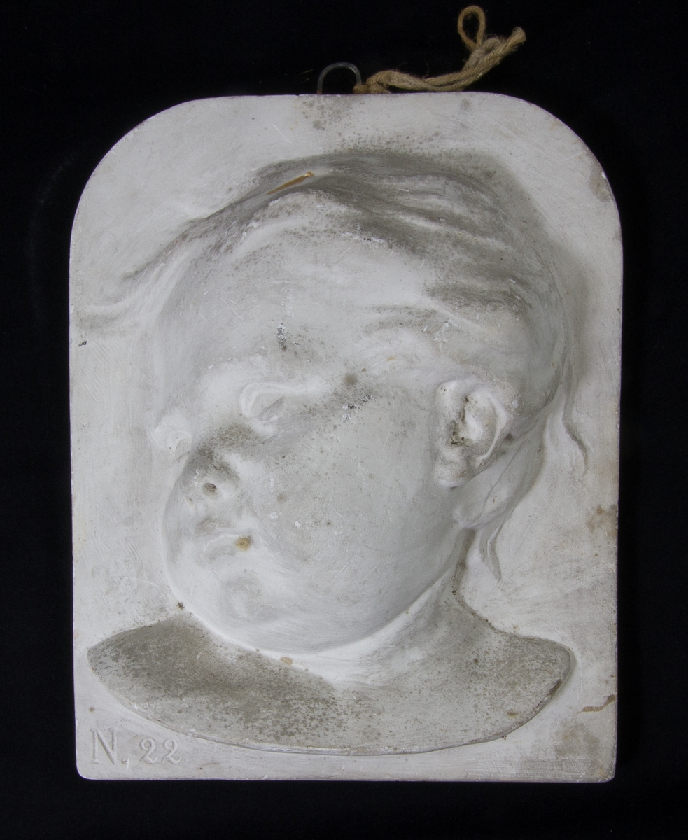 Porträttmedaljong av vit gips föreställande ett barnhuvud i hög relief.