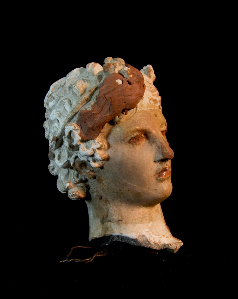 Studiemodell av gips föreställande ett kvinnohuvud. Håret, ögonen med mera förstärkt med plastelina av Julius Kronberg samt målad i rödbrunt. Sekundär ögla av järntråd för upphängning.