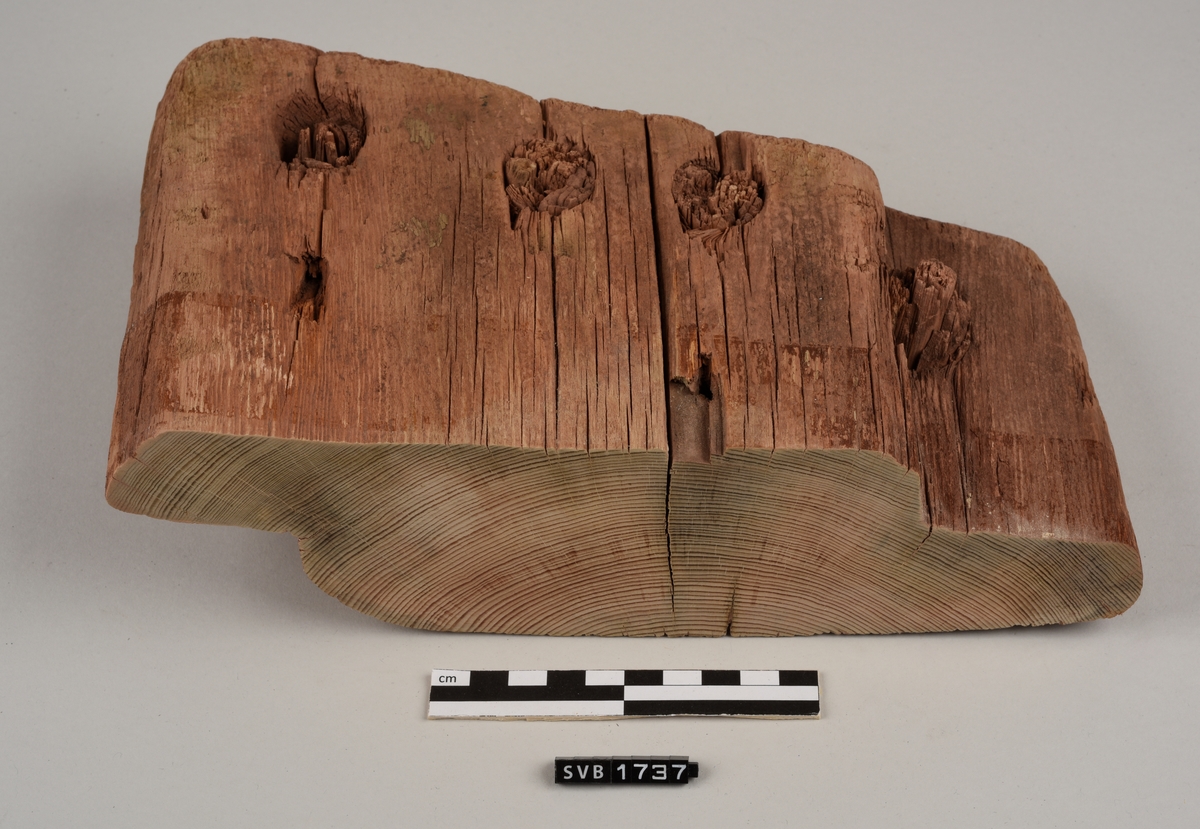 Del av bord til sydd båt. Det er hull med treplugger og sømhull med søm av røtter. Trematerialet har en rødlig farge. Antakelig på grunn av fargen på sanden der den var funnet.