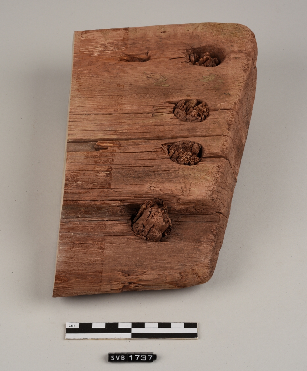 Del av bord til sydd båt. Det er hull med treplugger og sømhull med søm av røtter. Trematerialet har en rødlig farge. Antakelig på grunn av fargen på sanden der den var funnet.