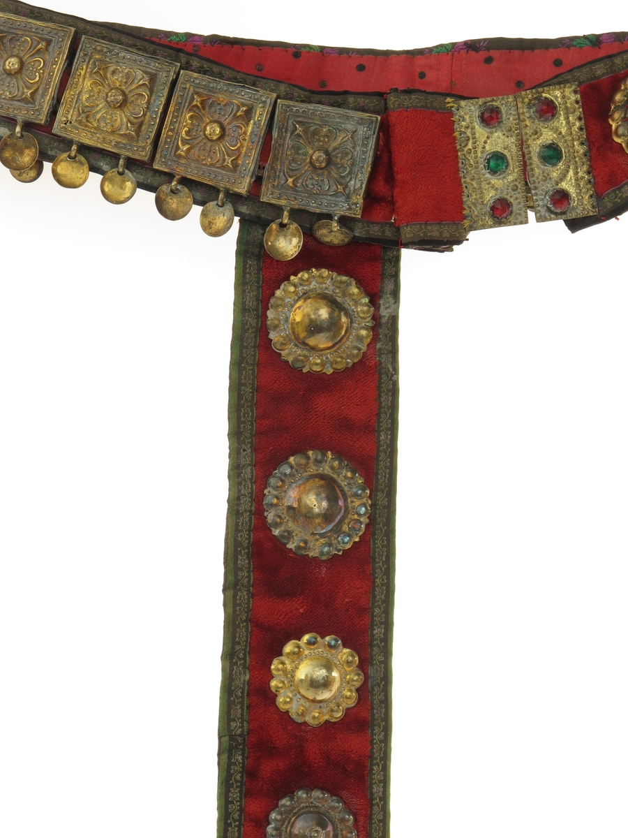 Belte med geheng. Av rød plysj, kantet med smal sort silkebord med små blomsterranker i grønt og fiolett. Beltet besatt med 16 kvadratiske utstansede forgylte sølvplater, hver med to nedhengende løv.  Flatene har rosettornamentikk.  Hektelås av forgylt blikk, de to deler hver med tre glasstener i rødt og grønt.  Ved siden av låsen på den ene side to runde forgylte beslag, utstanset arbeide, med ett løv i sentrum.  Festet til beltet et langt nedhengende geheng av samme materiale, besatt med ni runde utstansede forgylte beslag av forskjellig utførelse og størrelse.  Nederst beslått med avrundet endebeslag, forgylt, med utstanset ornamentikk på oversiden, et ansikt i midten.  Nederst tre løv. Beslaget har gullsmedmerker på baksiden (se Påført tekst) 
Beltet er foret med rødt bomullsstoff med sorte prikker.  Gehenget foret med sort silkebånd, blomsterranke i rosa, og tunget rosa kant.