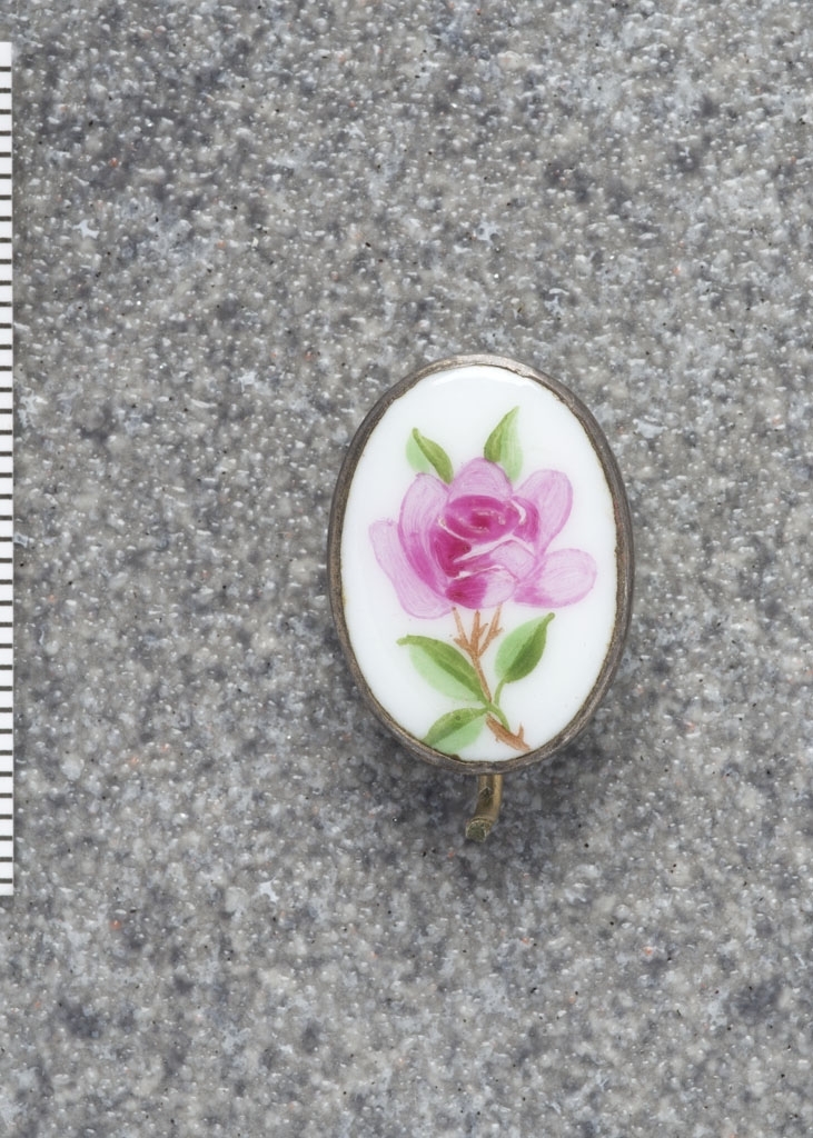 En brosje i emalje (antatt, muligens porselen). Oval form. På hvit bakgrunn en rosa rose med blader og stilk. Bak en krokete nål. Nålen er brukket.
