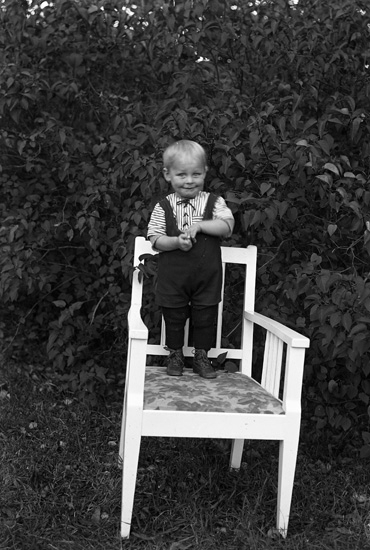 En liten pojke står på en vit karmstol.
Bakom honom finns lite lövträd