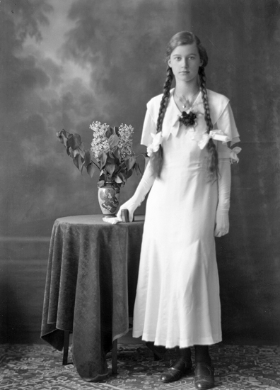 Foto av en kvinnlig konfirmand i konfirmationsklöänning. Hon står vid ett bord och håller i en psalmbok.
Helfigur, en face. Ateljéfoto.
